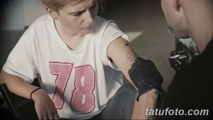 Фото тату: мастер наносит контур татуировки кота на плечо девушки. Виден эскиз рисунка и рука мастера с тату-машинкой.